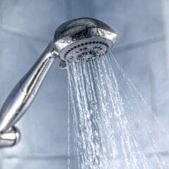 A silver showerhead spraying water in a bathroom, showcasing the artistry of bathroom installations. The water streams downwards against light blue tiled walls, creating a tranquil scene.