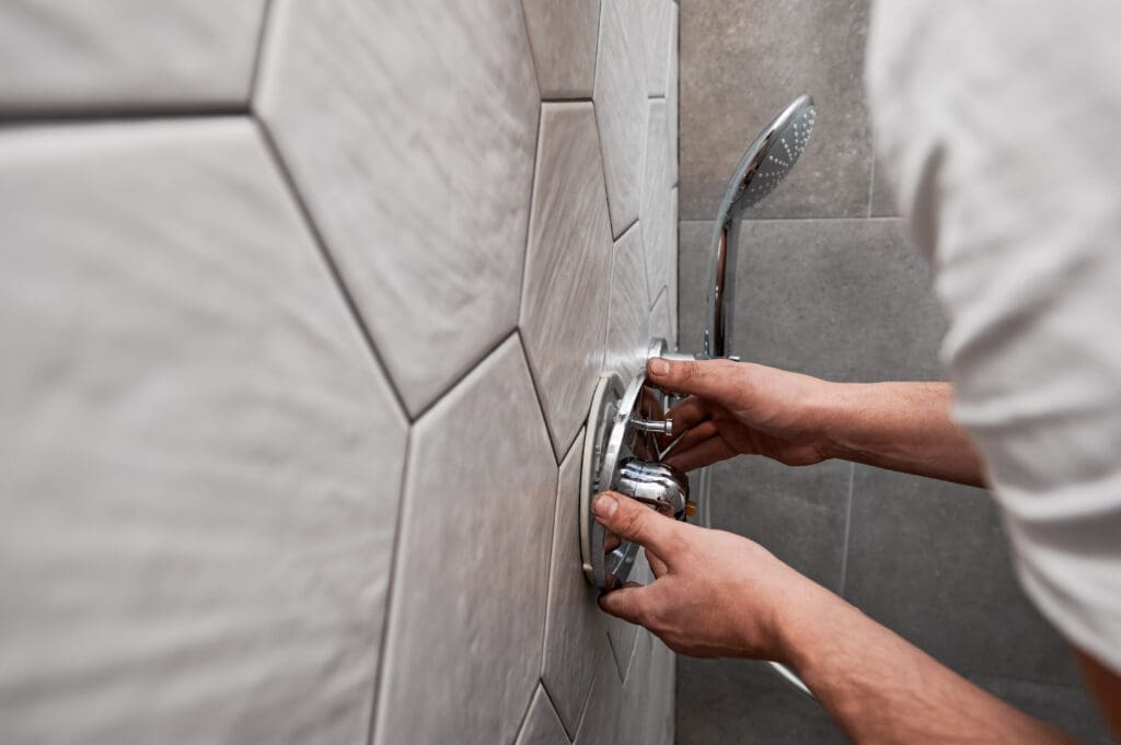 Close up of man standing by the wall with ceramic tile and installing shower faucet with metal handle in apartment. Male plumber working on bathroom renovation at home. Plumbing works concept.