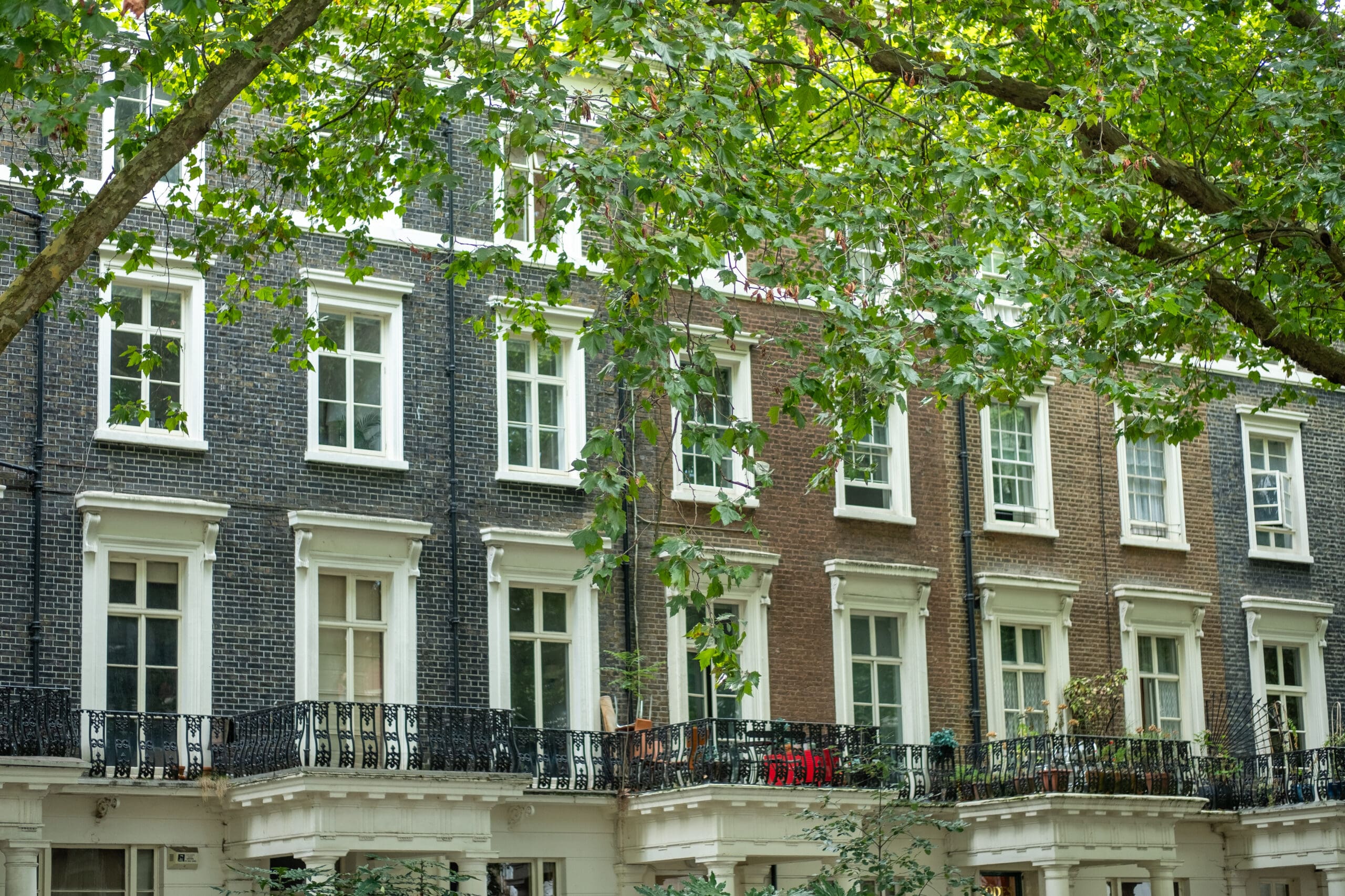 A row of charming city townhouses with brick facades features tall windows and ornate wrought iron balconies. Lush green trees extend in front, partially obscuring the view of these homes, known for their top-notch bathroom installations that blend style with modern convenience.