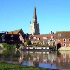 Scenic view of a historic riverside town with a prominent church spire. Brick and stone buildings line the river, where a moored narrowboat reflects in the calm water. This picturesque setting is perfect for exploring local property services or taking care of essential plumbing repairs under the clear blue sky.