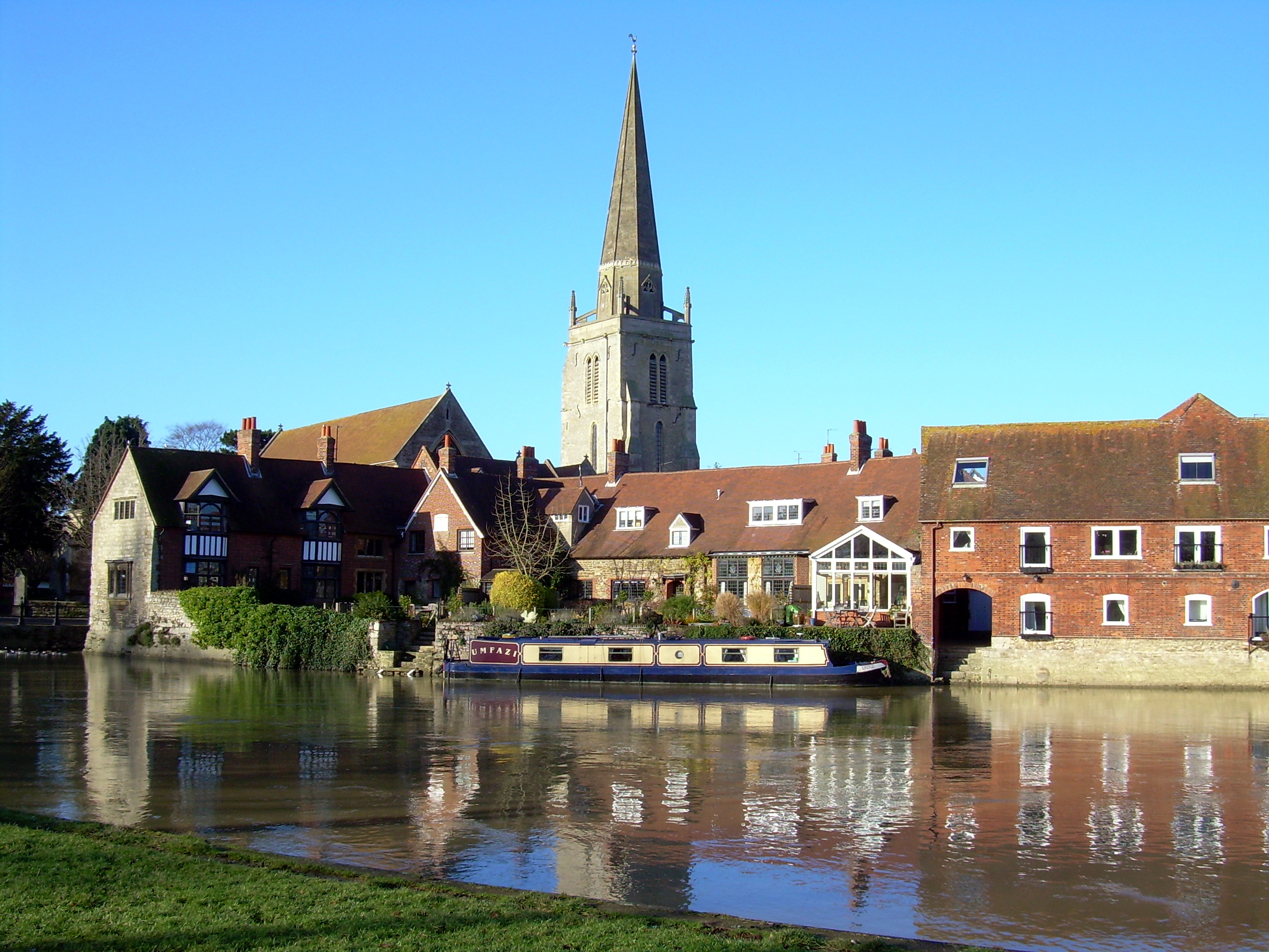 Scenic view of a historic riverside town with a prominent church spire. Brick and stone buildings line the river, where a moored narrowboat reflects in the calm water. This picturesque setting is perfect for exploring local property services or taking care of essential plumbing repairs under the clear blue sky.