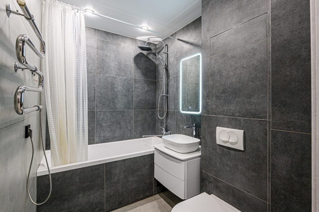 Modern bathroom with dark gray tiled walls and a white bathtub. It features a wall-mounted sink with a backlit mirror above, a rainfall showerhead, and a toilet. A sleek radiator adds warmth beside the curtain that partially encloses the bathtub area.