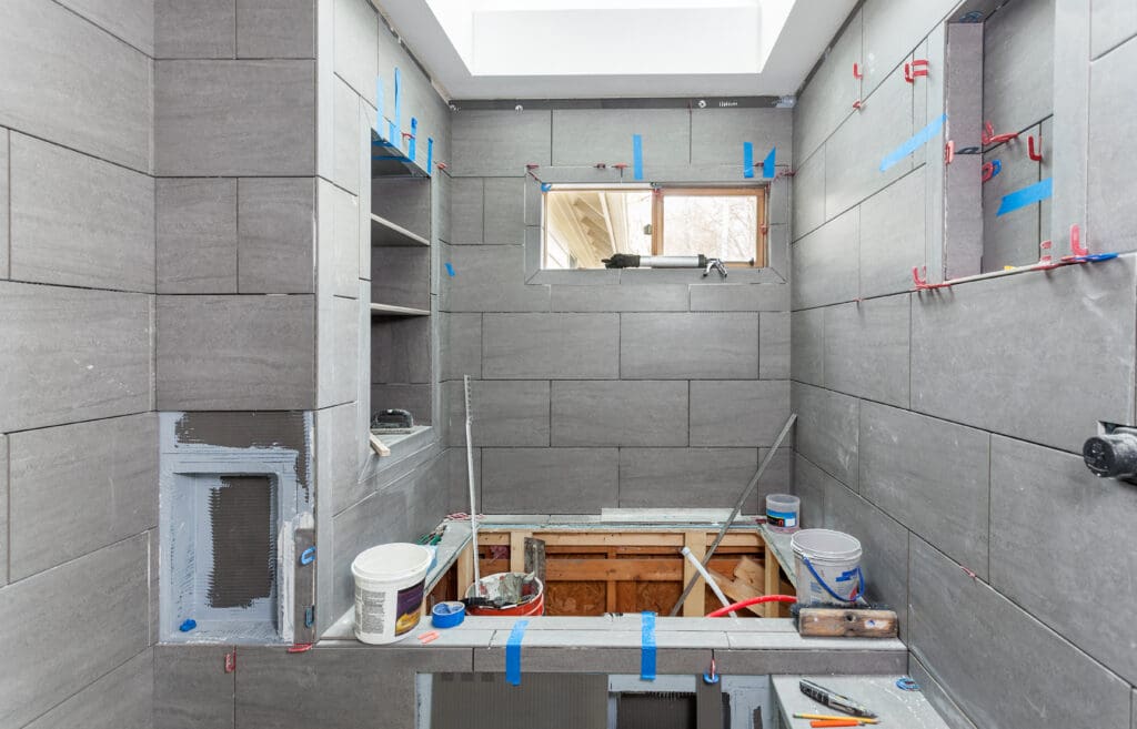 A bathroom under renovation with gray tiles partially installed and a plumber's tools scattered around. Paint cans and construction materials lie about, while shelves are in progress. Blue tape marks the walls, and a small window above lets in natural light, highlighting pending plumbing repairs.