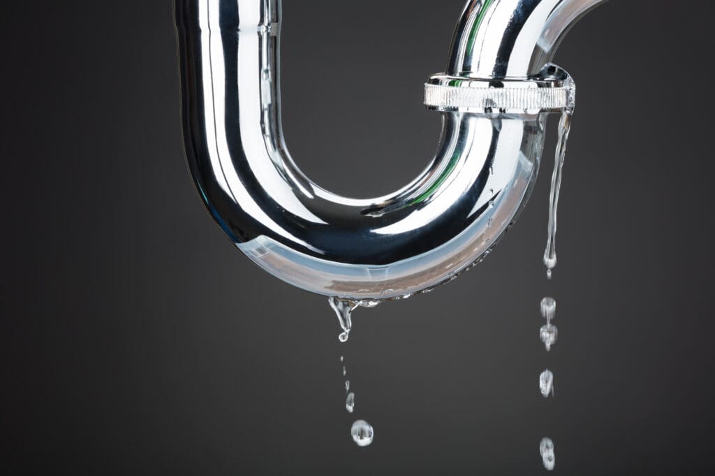 Close-up of a leaking metal pipe with water dripping from the underside, reminiscent of plumbing repairs in need. The dark background highlights the shiny surface and droplets.