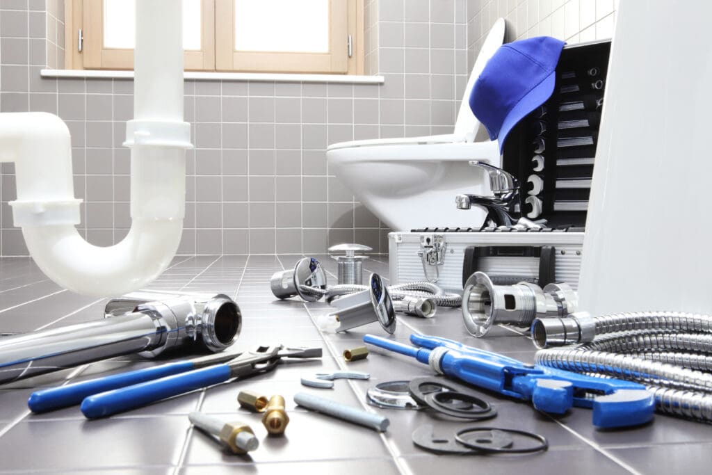 A bathroom scene showcases a plumber's workspace, with a toilet and toolbox surrounded by essential plumbing tools on the gray tiled floor. Wrenches and fittings are scattered around, alongside a blue cap atop the toolbox. The square-tiled walls feature a window in the background, highlighting ongoing bathroom installations.