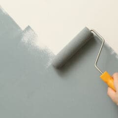 A hand with a yellow handle paint roller applies light gray paint to a wall, partially covering a white surface, hinting at a home improvement project in progress. Nearby radiators await the final touches for enhanced style and warmth.