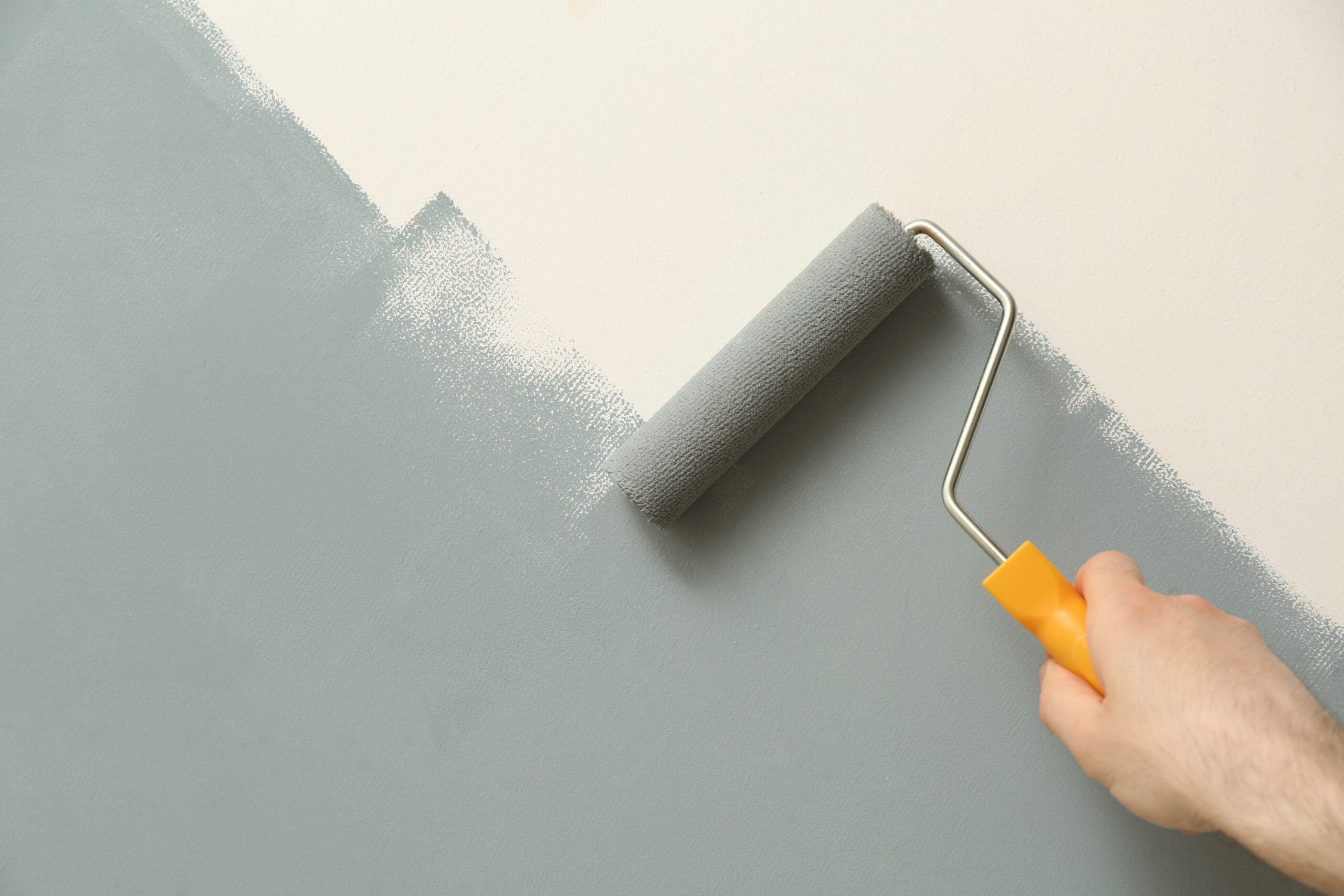 A hand with a yellow handle paint roller applies light gray paint to a wall, partially covering a white surface, hinting at a home improvement project in progress. Nearby radiators await the final touches for enhanced style and warmth.