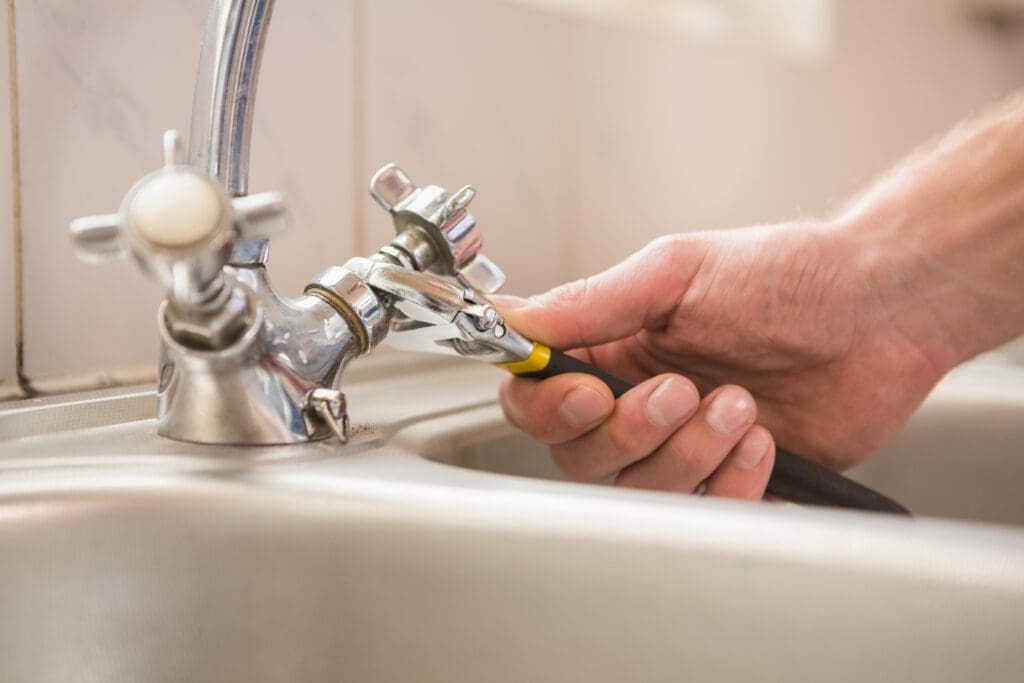 A person uses a wrench to tighten the fitting on a silver kitchen faucet, showcasing their knack for property services. The sink and tiled backsplash are visible in the background, hinting at bathroom installations or similar repair work.