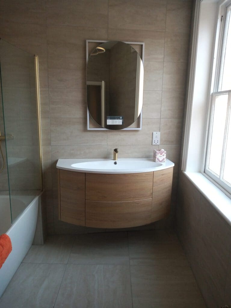 A modern bathroom showcases a wooden vanity with a white countertop and gold faucet. Above it, an oval mirror enhances the tiled wall. To the right, natural light streams through a window, illuminating the pristine layout of professional bathroom installations and adding charm to the visible bathtub on the left.