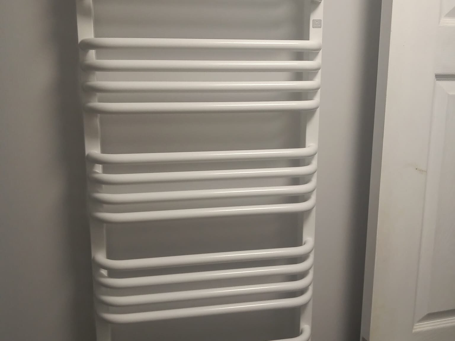 A white, wall-mounted towel radiator with multiple horizontal bars stands elegantly next to a closed white door. It exemplifies modern bathroom installations, expertly designed for heating and drying towels with ease.